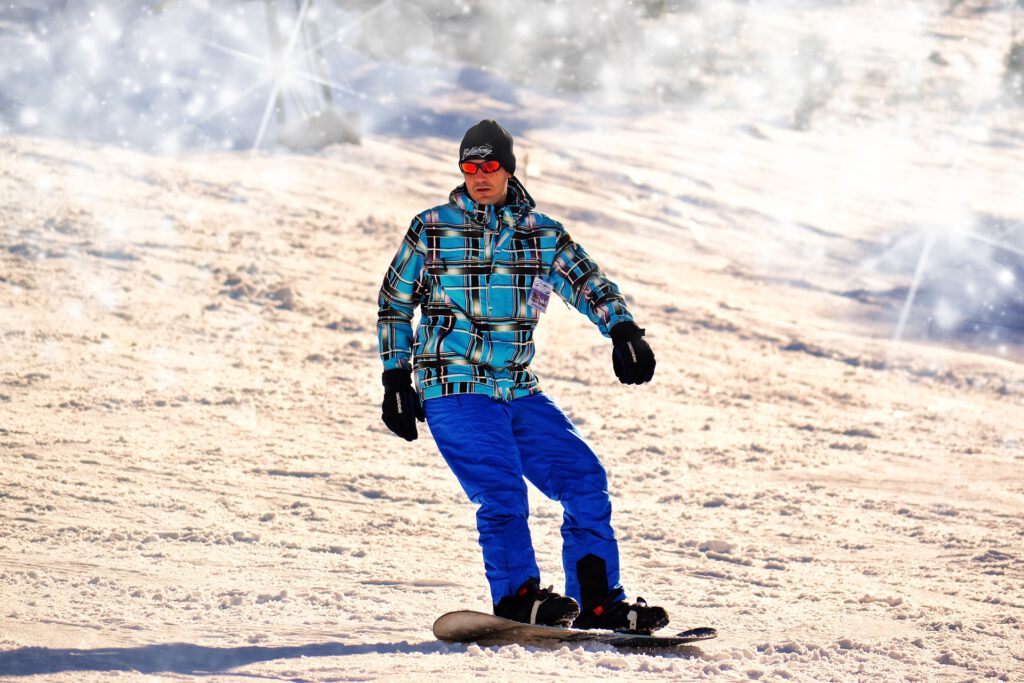 Mann im Schnee mit Snowboard, trägt Wollmütze, Sonnenbrille, winddichte Jacke und Hose und Handschuhe.
